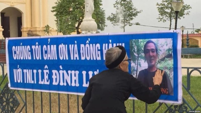 Thân mẫu nhà hoạt động Lê Đình Lượng chạm vào hình con mình trên một banner treo phía trước nhà thờ gần tòa án Vinh, 18 tháng 10, 2018. Ảnh: Facebook Nguyen Xoan.