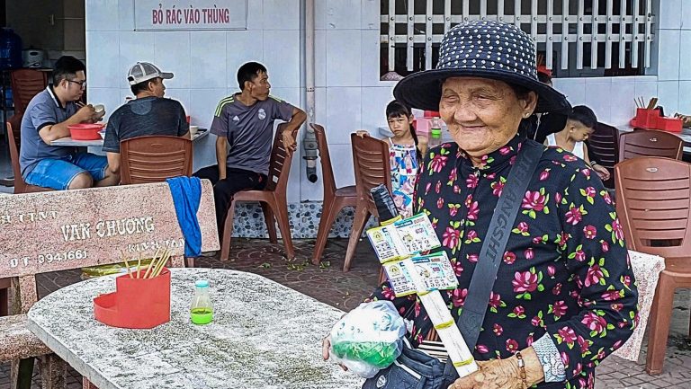 Một bà bán vé số dạo ở tỉnh Kiên Giang. Thu nhập từ bán vé số có thể đủ để trang trải chi phí hàng ngày. Cứ mỗi vé số bán ra, người bán lấy lời 1000 đồng. Một người bán chuyên nghiệp thường bán khoảng 200 vé mỗi ngày, thu chừng 200.000 đồng. Người bán nghiệp dư chỉ thu chừng 50.000 đồng mỗi ngày. Nói chung, thu nhập này đủ để họ sống qua ngày. Ảnh: Báo Tuổi Trẻ