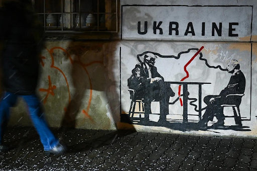Một bức tranh tường mang tên “Bị chia cắt bởi giới tài phiệt” của nghệ sĩ đường phố Na Uy ẩn danh Toddel khắc họa Tổng thống Mỹ Donald Trump và Tổng thống Nga Vladimir Putin ngồi bên bàn cạnh tấm bản đồ Ukraine bị chia cắt. Tác phẩm xuất hiện trên một bức tường ở Krakow, Ba Lan, ngày 29 tháng 1. Ảnh: Artur Widak/ Getty Images